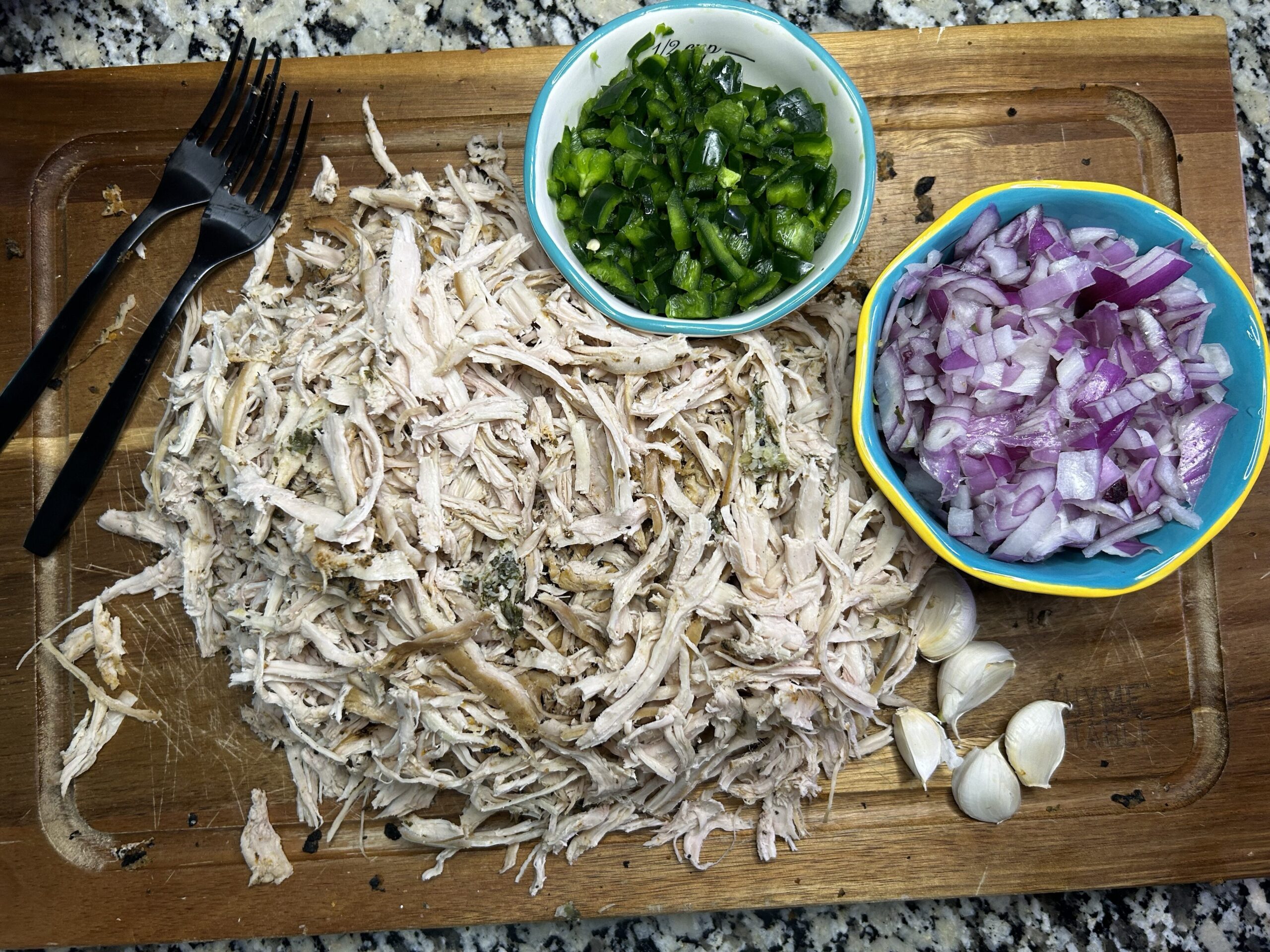 Shredded Turkey on a cutting board with a bowl on diced jalapeño and poblano peppers, a bowl of diced red onion, and garlic cloves