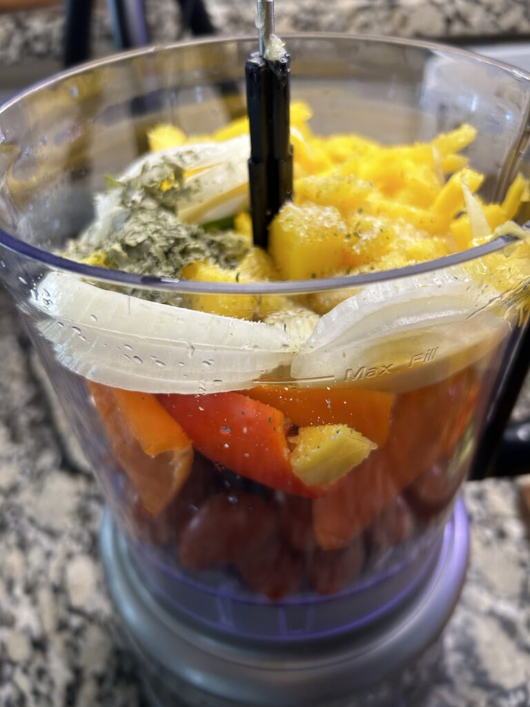 Food processor filled with sliced mango, onion, peppers, tomatoes, and seasonings.