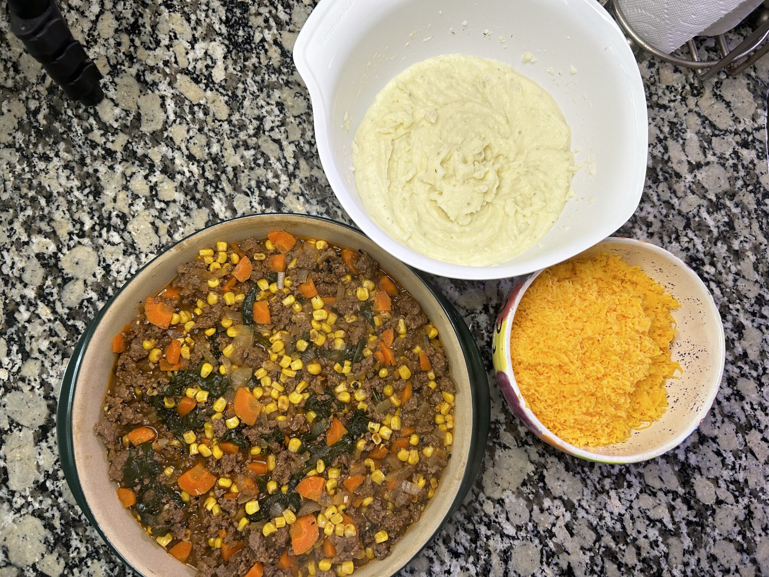 bowl of mashed potatoes, bowl of shredded cheddar cheese, and a dish filled with browned ground beef, sauteed spinach, carrots, onion, and corn
