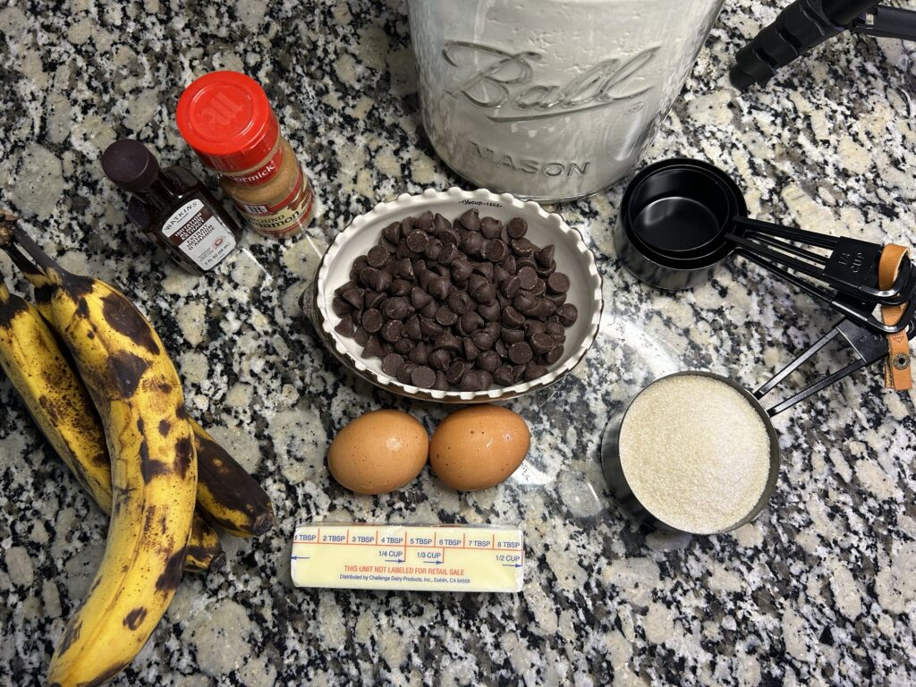 ripe bananas, stick of butter, 2 eggs, sugar, chocolate chips, flour, cinnamon, and vanilla extract on counter